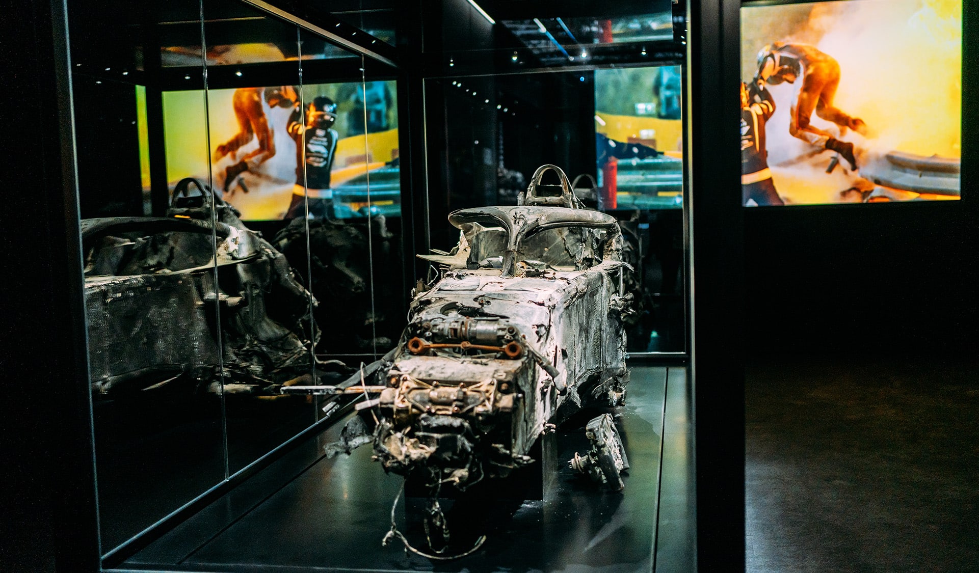 The charred chassis of Romain Grosjean's Formula 1 car from the Bahrain GP is displayed within a glass case. Screens in the background show images of the crash.