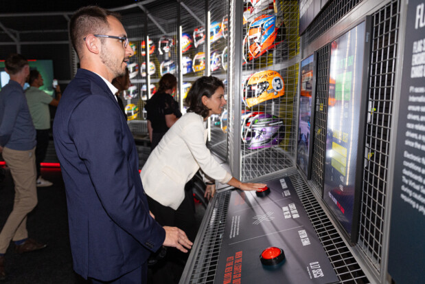 People interact with a multimedia station at the Formula 1 exhibition, pressing buttons and consulting screens, with a collection of drivers' helmets displayed in the background.