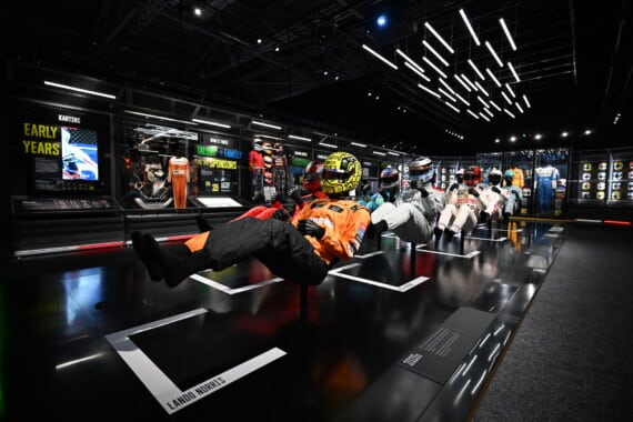 Mannequins in driving positions, wearing Formula 1 helmets and overalls, are suspended before panels detailing 'Early Years' and featuring Lando Norris's name, within a futuristic, brightly lit room that showcases the evolution of Formula 1 drivers.