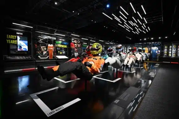 Mannequins in driving positions, wearing Formula 1 helmets and overalls, are suspended before panels detailing 'Early Years' and featuring Lando Norris's name, within a futuristic, brightly lit room that showcases the evolution of Formula 1 drivers.