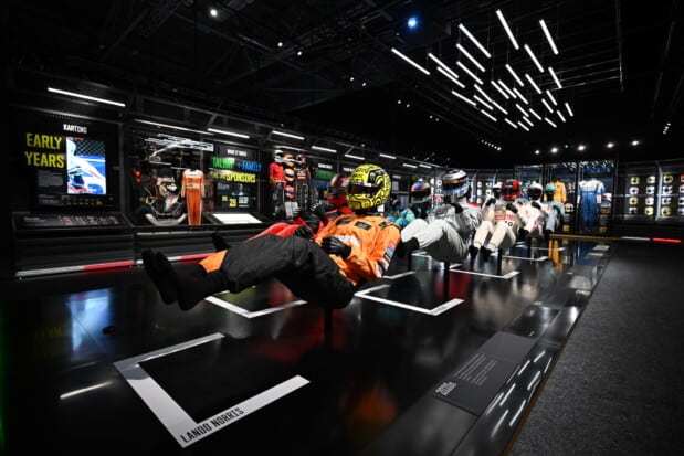 Mannequins in driving positions, wearing Formula 1 helmets and overalls, are suspended before panels detailing 'Early Years' and featuring Lando Norris's name, within a futuristic, brightly lit room that showcases the evolution of Formula 1 drivers.