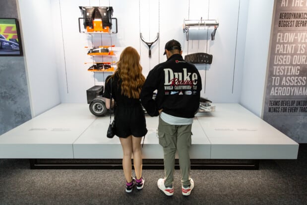 Visitors stand with their backs to the camera, observing a display of motorsport parts and components. A nearby wall explains the aerodynamic tests conducted by teams to enhance the performance of Formula 1 cars.
