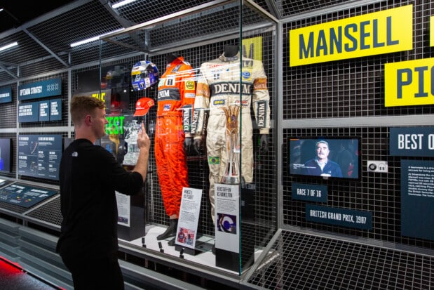 A visitor stands before a latticed showcase displaying two 'Denim' branded Formula 1 racing suits (one orange and one light), helmets, caps, and large yellow signs bearing the surnames 'Mansell' and 'Piquet'. To the right, a screen shows information about the 1987 British Grand Prix, race 7 of 16, as the visitor examines these historical motorsport artifacts.