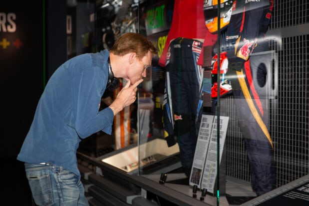 A man wearing a blue shirt leans forward, closely observing racing suits and a Red Bull-branded helmet within a showcase