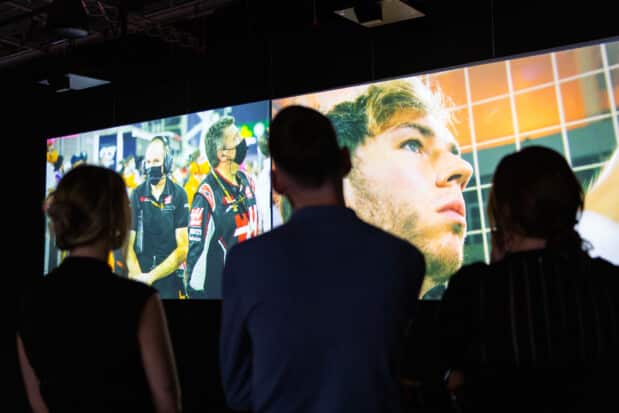 A group of people, with their backs turned, are viewing Formula 1 images on two screens