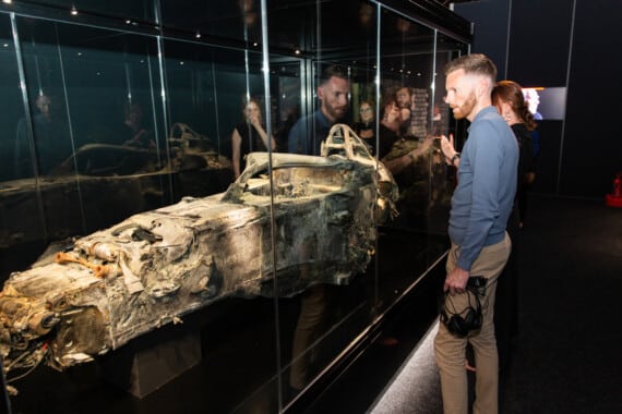 A man with headphones in hand examines the charred chassis of Romain Grosjean's Formula 1 car inside a glass case.