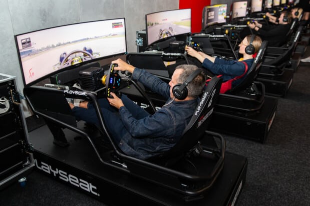 A row of racing simulators, featuring panoramic screens and Playseat seats, allows visitors to drive virtual vehicles with steering wheels and headsets, providing an immersive Grand Prix experience