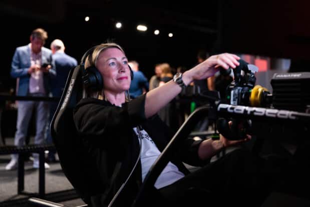 A woman with a headset sits in a Playseat simulator, driving with a Fanatec steering wheel, immersed in the driving experience as others converse in the background