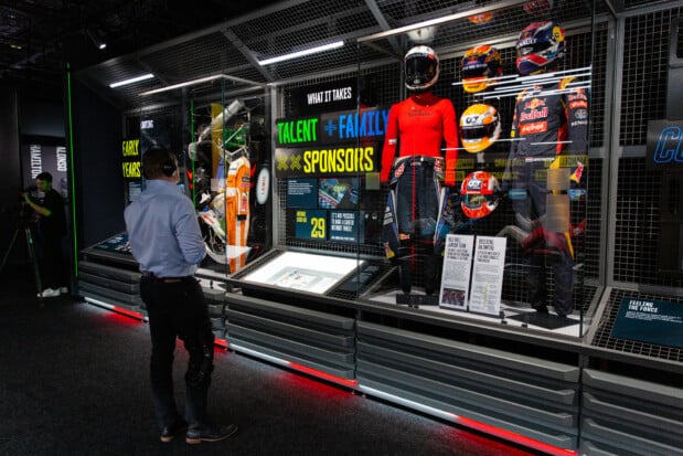 A visitor with a headset stands before a showcase displaying racing suits, helmets with Red Bull logos, and information panels titled 'Talent + Family + Sponsors' and 'Early Years', highlighting key factors in a Formula 1 driver's career